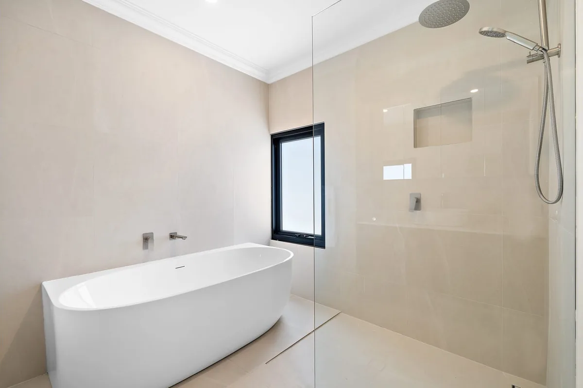 Freestanding white bath beside a glazed shower and warm neutral wall tiles.